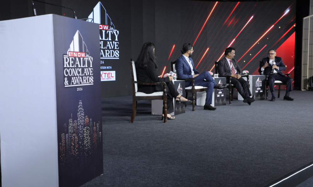 Vineet Nanda speaking at an industry event with a modern Indian skyline, residential towers and smart city infrastructure in the background, representing the future of India's real estate market.