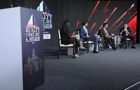 Vineet Nanda speaking at an industry event with a modern Indian skyline, residential towers and smart city infrastructure in the background, representing the future of India's real estate market.